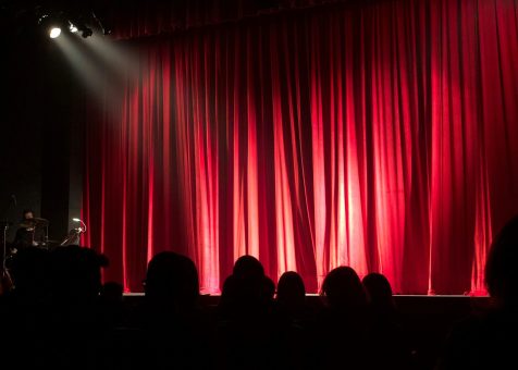 Dimly lit theater stage with red curtains and audience silhouettes under spotlights.